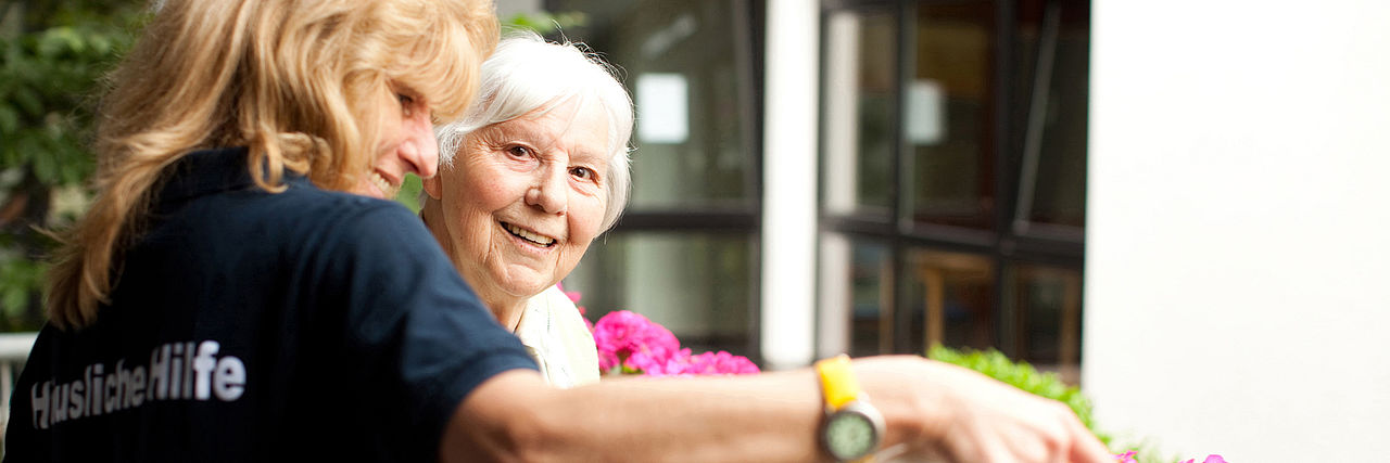 Hauswirtschaftliche Hilfen des DRK. Eine lachende Seniorin mit einer DRK-Mitarbeiterin beim Blumen gießen.